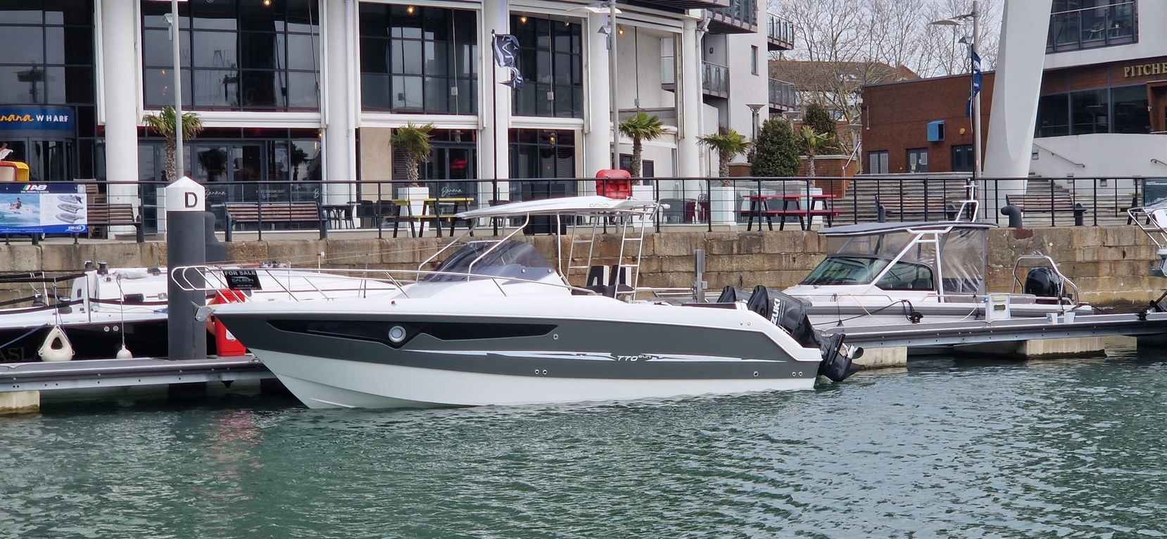 A sleek motorboat docked at a marina near a modern waterfront building with palm trees and seating areas.